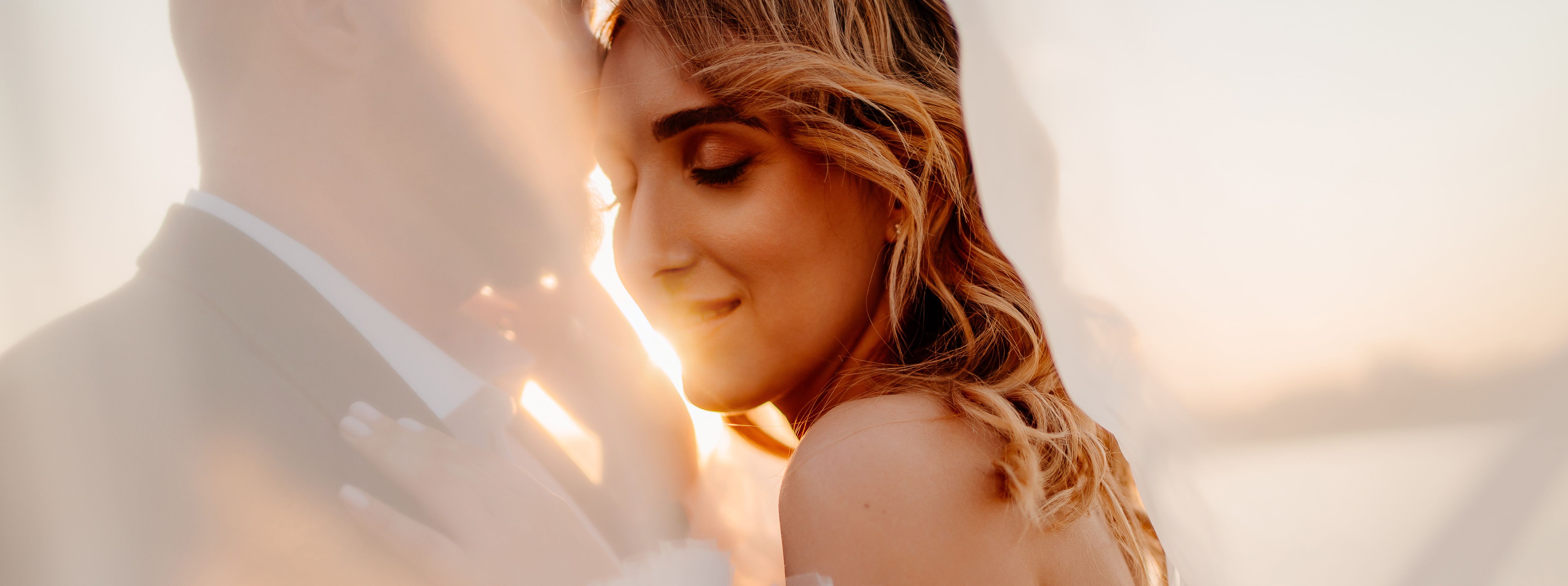 A bride smiles while being held by her groom, as they stand in front of a sunset on the water.