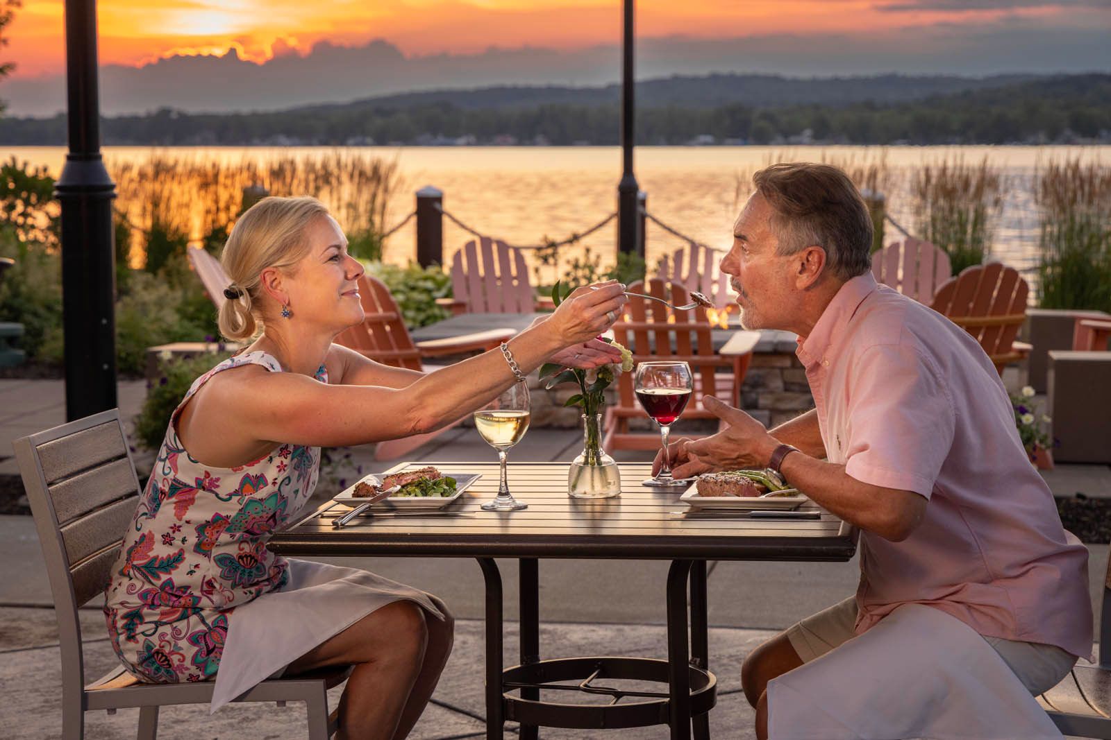A woman feeds her partner over the table as they dine outside on the patio in front of a beautiful sunset at the Chautauqua Harbor Hotel